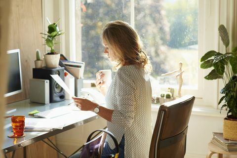 Γυναίκα πίνοντας καφέ και την ανάγνωση χαρτιά στο γραφείο σε ηλιόλουστο σπίτι γραφείο.