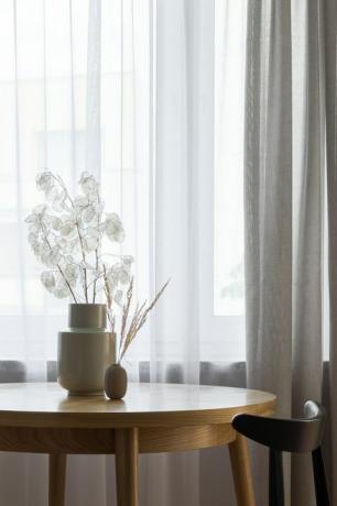 ωραίο δωμάτιο με στρογγυλό, ξύλινο τραπέζι φαγητού με φυσική διακόσμηση κοντά σε παράθυρο με λευκές και μπεζ κουρτίνες