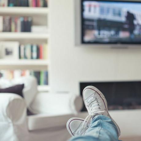 Εικόνα τζιν και αθλητικά παπούτσια, πόδια στον καναπέ, με τηλεόραση, τζάκι, καναπέ και βιβλιοθήκη στο παρασκήνιο.